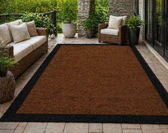 May include: A rectangular brown outdoor rug with a black border. The rug is placed on a stone patio, with a wicker sofa and chair, and potted plants in the background. The rug is a solid colour.