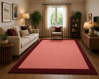 May include: A rectangular rug with a coral centre and a deep burgundy border. The rug is in a living room with a sofa, chairs, and a wooden side table. The room has natural light.
