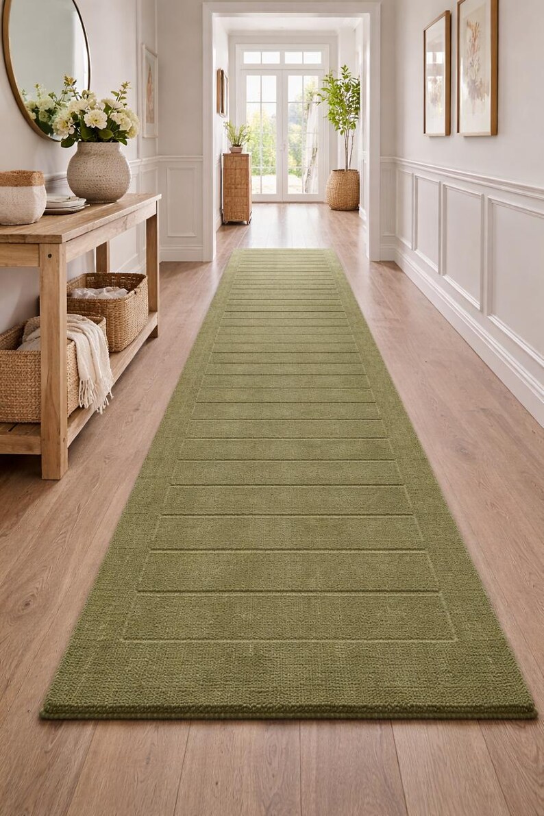 Puede incluir: Una alfombra larga de pasillo verde oliva con un dise&ntilde;o rectangular. La alfombra presenta una superficie texturizada con l&iacute;neas horizontales, creando un patr&oacute;n sutil. Est&aacute; colocada sobre un suelo de madera en un pasillo bien iluminado.