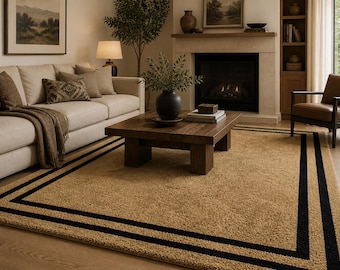 May include: A beige rug with a black border in a living room. The rug is under a wooden coffee table and in front of a fireplace. A sofa and armchair are also visible.