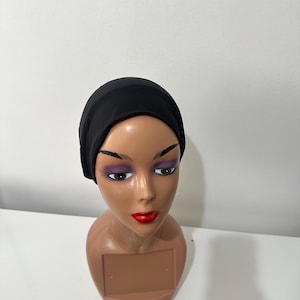May include: A black head covering is displayed on a mannequin head. The mannequin has brown skin, red lipstick, and purple eyeshadow. The head covering is made of a soft, opaque material.