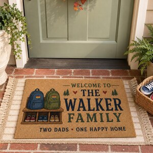 May include: A rectangular tan coir doormat featuring a graphic of two backpacks on a shelf above two pairs of shoes. The text reads: WELCOME TO THE WALKER FAMILY, TWO DADS ONE HAPPY HOME. The design includes small pine trees and red heart icons.