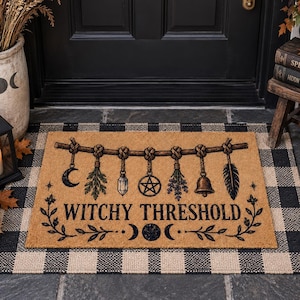 May include: A rectangular tan coir doormat featuring a black printed design of a horizontal branch with hanging charms including a crescent moon, herbs, a crystal, a pentagram, a bell, and a feather. The text WITCHY THRESHOLD is centered below.