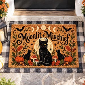 Peut inclure: Un paillasson rectangulaire en fibre de coco avec le texte Moonlit Mischief en lettres noires. Le motif comprend un chat noir central, deux petits chats noirs, des citrouilles orange, des champignons rouges, des glands, du feuillage et des lunes.