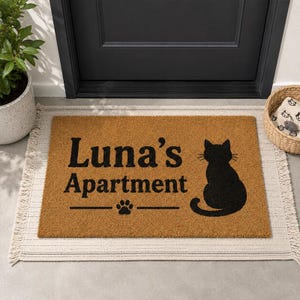 Peut inclure: Un paillasson rectangulaire en fibre de coco marron avec le texte noir Luna's Apartment, une silhouette noire de chat assis et une petite icône de patte noire centrée sous le texte, le tout sur une base en tissu tissé clair.