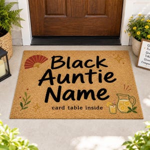 May include: A rectangular doormat with a tan background. The mat features the words "Black Auntie Name" in large black letters. Decorative elements include a red fan, a pitcher of lemonade, and the text "card table inside."