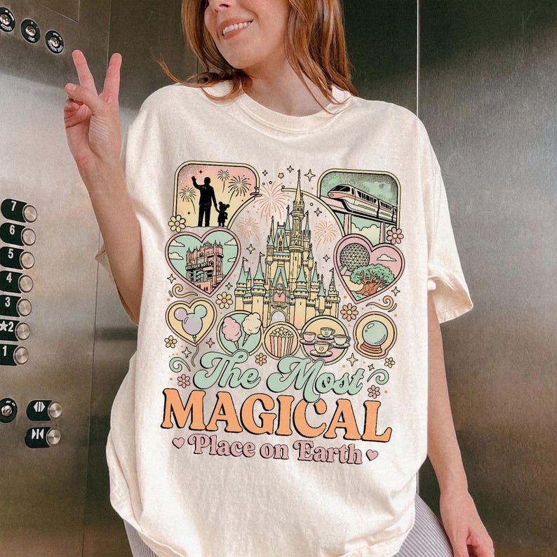 May include: Cream-colored t-shirt with a colorful graphic design featuring a castle, train, and other theme park icons. The text "The Most Magical Place on Earth" is printed in a whimsical font.