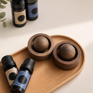 May include: Two dark brown wooden circular holders, each containing a smooth wooden sphere, are placed on a light wood oval tray. Four small black glass bottles with labels reading Essential Oil, Pure & Natural, and Aromatic Grade are included.