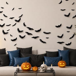 May include: A collection of black bat-shaped wall decals arranged in a swarm pattern above a sofa. The sofa features black and blue pillows, two glowing orange jack-o'-lanterns, a white skull, and two small candles on a black coffee table.