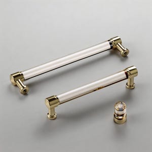 Könnte beinhalten: Zwei Schrankgriffe mit einem transparenten, zylindrischen Mittelteil und goldfarbenen Metallenden. Ein Griff ist länger als der andere. Ein kleiner, passender Knopf ist ebenfalls vorhanden, alles auf grauem Hintergrund.