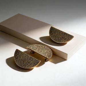 Può includere: Tre maniglie per mobili a forma di semicerchio color ottone con un motivo lineare strutturato. Due maniglie sono posizionate su una superficie bianca, mentre la terza è su un blocco rettangolare beige. Le maniglie hanno piccoli piedini arrotondati.