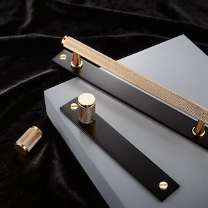 May include: Close-up of several cabinet hardware pieces. The hardware includes a long, textured gold handle, a gold knob, and black rectangular plates with gold accents. The pieces are set against a gray and black background.