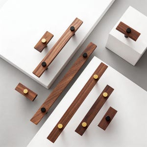 May include: A collection of wooden cabinet pulls in various sizes. The pulls are a rich brown color with contrasting black and gold-colored hardware. The hardware is circular and adds a modern touch to the design. The pulls are arranged on a white surface.