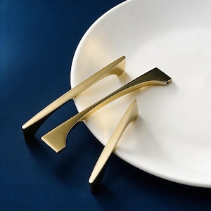 May include: A close-up shot of three gold-colored cabinet pulls arranged on a white plate against a dark blue background. The pulls have a sleek, modern design with a rectangular base and a tapered handle. The lighting highlights the metallic finish.