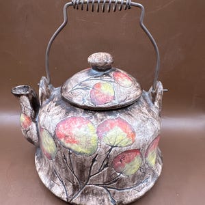 May include: A ceramic teapot with a decorative leaf pattern in red, yellow, and green on a brown background. The teapot has a metal handle and spout, and a matching lid with a knob.