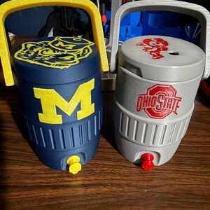 May include: Two small beverage coolers with handles and spigots. The left cooler is navy blue with a yellow block M and a mascot logo on the lid. The right cooler is gray with a red Ohio State logo on the front and a red mascot logo on the lid.