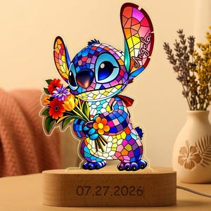 May include: A decorative acrylic lamp featuring a colorful, stained-glass style illustration of the character Stitch holding a bouquet of flowers. The name Angela is printed on one ear. The lamp sits on a wooden base engraved with 07.27.2026.