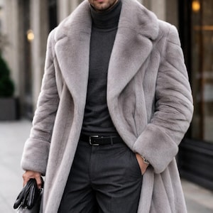 Könnte beinhalten: Ein langer, hellgrauer Kunstpelzmantel mit breitem Revers, getragen über einem dunkelgrauen Rollkragenpullover und einer dunkelgrauen Hose mit schwarzem Gürtel. Das Outfit wird durch schwarze Lederhandschuhe und eine schwarze Aktentasche ergänzt.