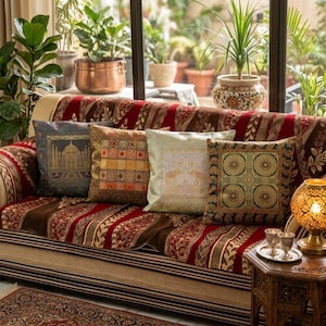 Op de afbeelding: Een bank met kussens met patronen in blauw, goud en bruin. De bank heeft een rood en bruin patroon. Een kleine houten tafel met een decoratieve lamp en twee glazen staat naast de bank. Planten zijn zichtbaar op de achtergrond.