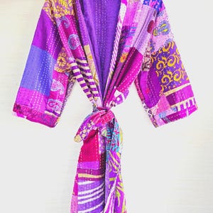 May include: A patchwork robe in shades of purple, pink, and yellow, hanging on a wooden hanger. The robe features a variety of patterned fabric squares, stitched together with white thread. The robe is tied at the waist with a matching fabric belt.