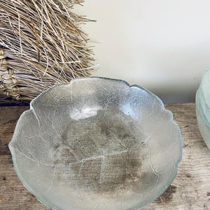 Puede incluir: Un cuenco de vidrio transparente con forma de hoja, con detalles de vetas grabados en la superficie. El cuenco está sobre una superficie de madera, con un elemento de paja tejida en el fondo. El diseño evoca una estética natural y orgánica.