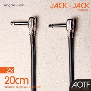 Può includere: Due cavi audio neri con connettori argentati, ciascuno di 20 cm di lunghezza, sono mostrati su uno sfondo beige chiaro. Il testo "JACK - JACK pancake" è arancione. L'immagine include anche il testo "mogami + rean" e "AOTF handcrafted audio gears".