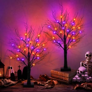 May include: Two decorative black leafless trees with illuminated branches featuring small purple and orange LED lights. The trees stand on square bases, with one placed on a stack of books, surrounded by miniature ghost and skull figurines.