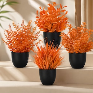 Pode incluir: Um conjunto de quatro plantas artificiais com folhagem laranja vibrante, cada uma num recipiente arredondado preto mate e liso. A coleção inclui várias formas de folhas, incluindo frondes de feto, folhas arredondadas e lâminas de erva.
