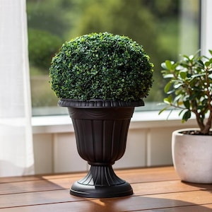May include: A black urn-shaped planter featuring a fluted base and rim, containing a dense, spherical topiary of small green leaves. The planter has a matte finish and a wide, flared opening that supports the rounded shape of the foliage.