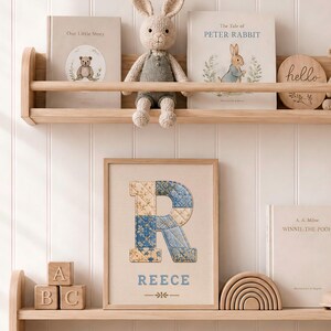 May include: A framed wall art print featuring a large letter R with a blue and beige quilted pattern, the name REECE in blue text, and a small floral icon. The print is displayed on a wooden shelf alongside a crocheted rabbit, books, and blocks.