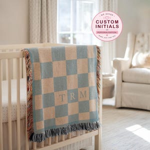 May include: A blue and beige checkered baby blanket with fringe, draped over a white cot. The blanket features the initials "TRM" embroidered in the centre. A pink circle graphic in the upper right corner reads "We offer custom initials or personalisation."