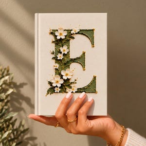 May include: A hardcover book with a cream-colored cover featuring a large, dark green letter E decorated with white flowers and green leaves. The floral design is textured and accented with gold lines, creating a detailed botanical illustration.