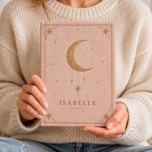 May include: A dusty pink hardcover journal featuring a gold foil crescent moon and star design on the front cover. The name ISABELLE is printed in gold lettering below the moon. The cover includes a thin gold border with star accents.