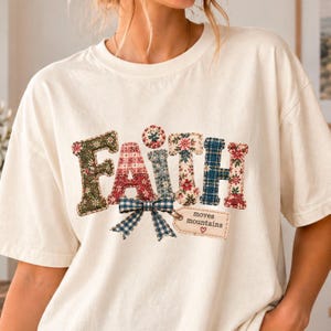 May include: An off-white short-sleeve t-shirt featuring the word FAITH in large, multi-patterned, quilt-style letters. A blue and white gingham bow sits below the letters, attached to a small rectangular tag that reads moves mountains with a heart.