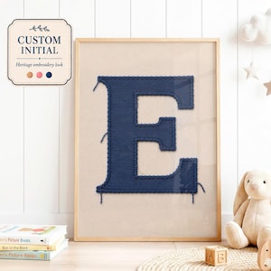 May include: A framed wall art print featuring a large, dark blue letter E with a textured embroidery-style appearance on a light beige background. The text CUSTOM INITIAL and Heritage embroidery look are visible in a small label at the top left.
