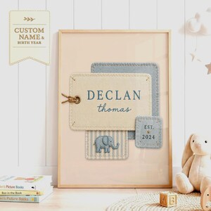 May include: A framed wall art print featuring layered fabric-style patches in cream and blue. The central cream patch displays the name DECLAN thomas in blue text. A smaller blue patch shows EST. 2024 with a star, and a striped patch features an elephant.