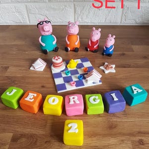 Pode incluir: Um conjunto de figuras coloridas de fondant, incluindo personagens de porcos, blocos de letras que soletram "GEORGIA" e um pequeno bolo. O conjunto está disposto sobre uma superfície de madeira, com o texto "SET 1" em vermelho.