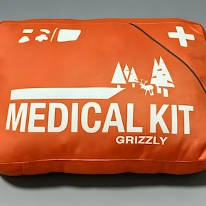 May include: A rectangular orange medical kit bag featuring white printed graphics including a cross symbol, a forest scene with a deer, and the text MEDICAL KIT GRIZZLY. The bag has a smooth, durable fabric construction with a diagonal line detail.
