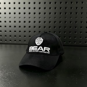Op de afbeelding: Een zwarte baseballpet met wit geborduurde tekst die BEAR en PRIVATE MILITARY CONTRACTOR leest, met een wit schildvormig logo boven de tekst dat het woord BEAR bevat. De pet heeft een gestructureerde kroon en een gebogen klep.