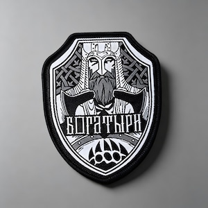 Könnte beinhalten: Ein schildförmiger Stickaufnäher mit einer schwarz-weißen Grafik eines bärtigen Kriegers mit Helm, der zwei Äxte hält. Das Design umfasst komplizierte Knotenmuster und den kyrillischen Text BOGATYRI über einem stilisierten Bärenpfotenabdruck.