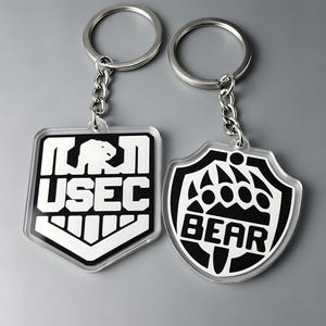 May include: Two acrylic keychains with silver metal rings and chains. The left keychain is a shield shape with a black background, white border, and the white text USEC above a graphic. The right keychain is a shield shape with a white background, black border, and the black text BEAR below a graphic.