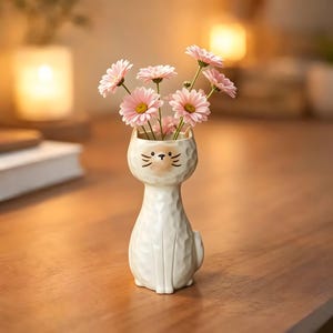 May include: A white ceramic cat-shaped vase with a textured surface, holding pink daisies with yellow centers. The vase has a painted face with whiskers and is sitting on a wooden surface.