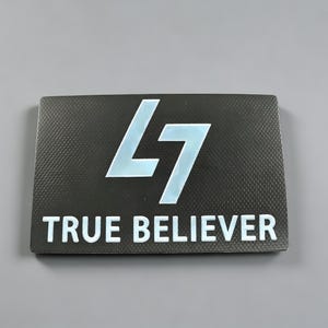 Könnte beinhalten: Eine rechteckige schwarze Plakette mit texturiertem Kohlefaser-Muster. In der Mitte befindet sich ein großes, stilisiertes hellblaues L7-Logo. Unter dem Logo ist der Text TRUE BELIEVER in fetten, weißen, serifenlosen Großbuchstaben aufgedruckt.