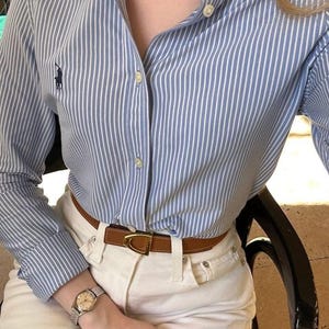 Op de afbeelding: Een lichtblauw en wit verticaal gestreept overhemd met een klein donkerblauw geborduurd logo op de borst, gecombineerd met een witte broek, een bruine leren riem met een gouden gesp, een parelketting en een zilverkleurig polshorloge.