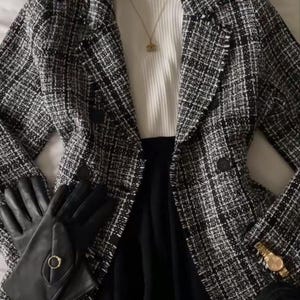 Op de afbeelding: Een zwart-witte tweed blazer met een dubbele rij knopen, gedragen over een witte geribbelde top met opstaande kraag. Accessoires zijn een zwarte haarband, een gouden ketting met hanger, zwarte leren handschoenen met een gouden ring, een gouden horloge en zwarte loafers.