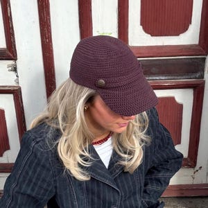 May include: A dark purple crocheted cap with a button detail. The cap has a visor and a textured appearance. The cap is a stylish accessory.