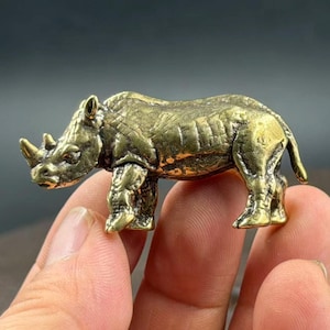 May include: A small, metallic brass figurine of a rhinoceros. The sculpture features a textured, weathered surface with visible folds and creases on the skin, two prominent horns on the snout, and a sturdy, four-legged stance in a side profile.