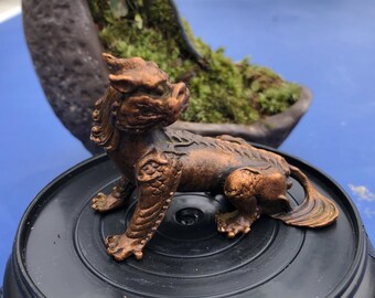 May include: A bronze-colored, detailed figurine of a mythical creature, possibly a guardian lion, with intricate carvings. The figurine is positioned on a black base, with a blurred background featuring a dark, oval-shaped planter with greenery.