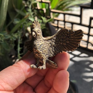 Könnte beinhalten: Detaillierte Metallfigur in Bronzeoptik, die einen Vogel mit ausgebreiteten Flügeln darstellt. Der Vogel hat einen Kamm auf dem Kopf und detaillierte Federn. Die Figur wird in einer Hand vor einem verschwommenen Hintergrund aus Grün gehalten.