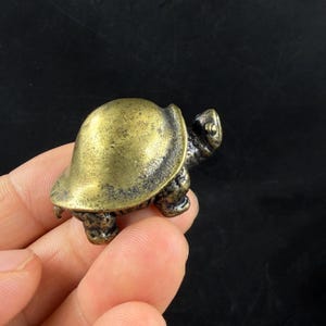 May include: A small, antique-style brass turtle figurine. The turtle has a detailed shell and textured skin, with a weathered appearance. The figurine is held in a hand, showcasing its size and craftsmanship. The turtle is a decorative item.
